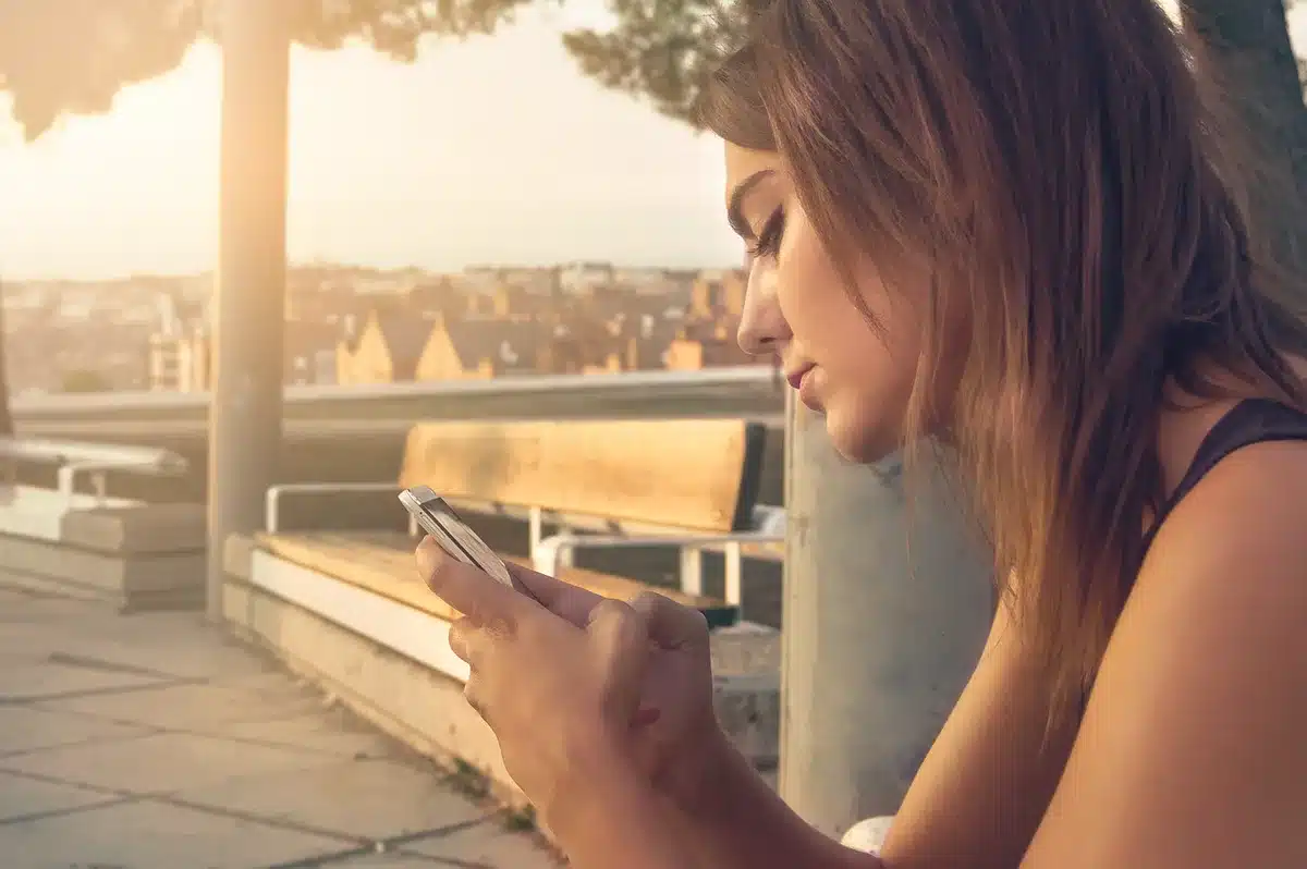 A close-up of an unbranded smartphone lying on a wooden table, screen off, showing a shadowed silhouette and a visible cooling vent on the side; anonymous hands reach for the device in soft focus