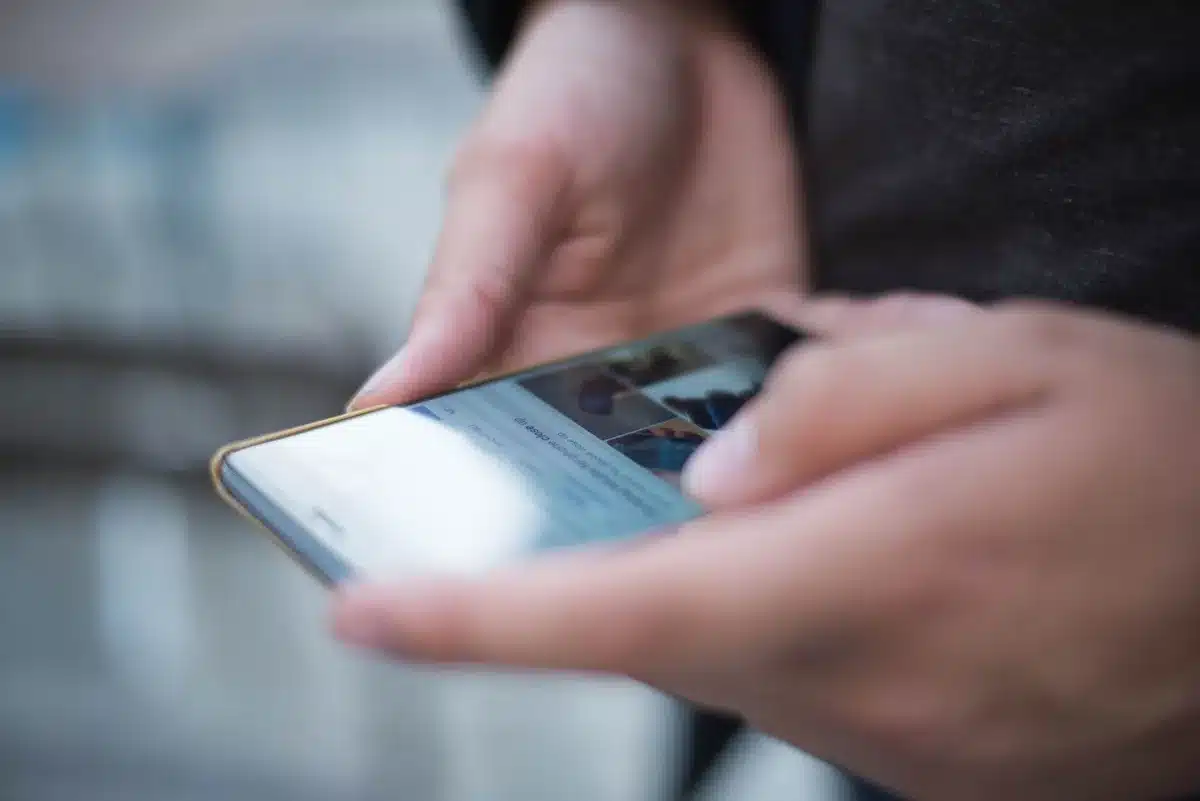Anonymous hands holding a phone with screen slightly angled away from surrounding blurred figures in a cafe, implying privacy in public; faces unrecognizable and no branding visible